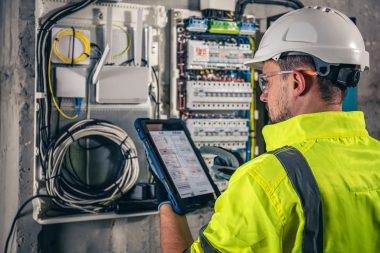 Man, an electrical technician working in a switchboard with fuses, uses a tablet. Formation Habilitation Électrique H0B0